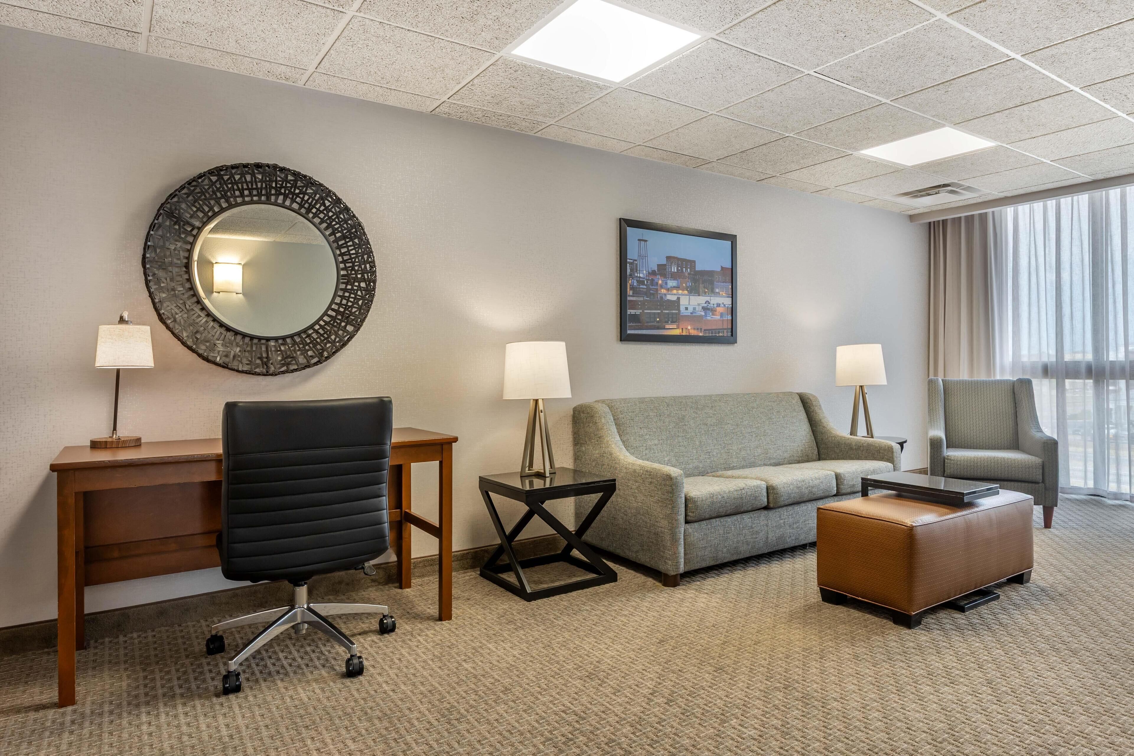 A hotel room with a desk, office chair, two lamps, a sofa, a chair, and a small ottoman.