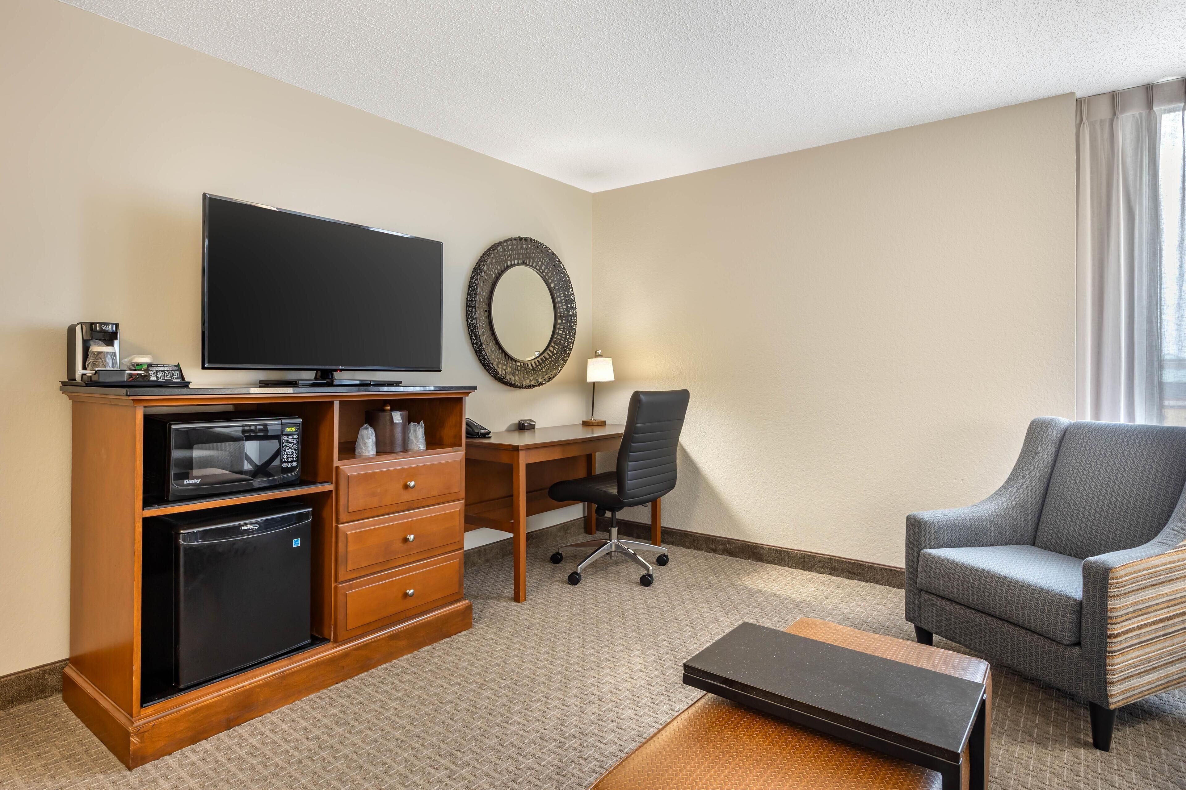 A hotel room with a flat-screen TV, a microwave, a refrigerator, a desk with a chair, a sofa, and a large mirror.
