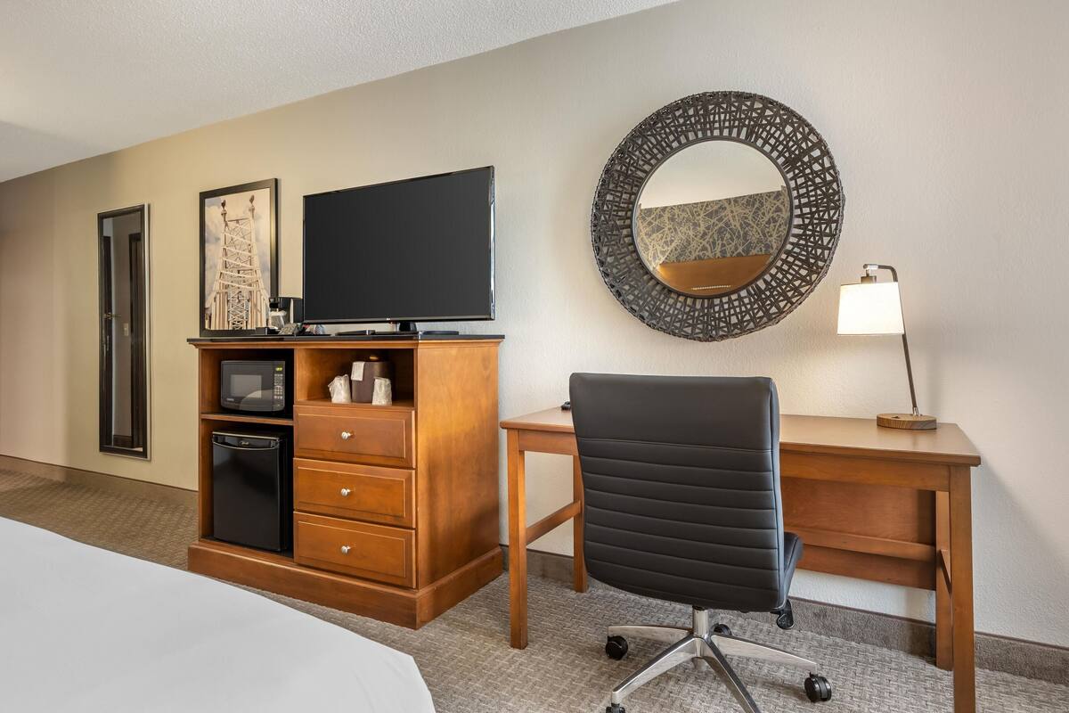 A hotel room with a bed, a wooden desk, a black office chair, a television, a microwave, a small cabinet, a decorative mirror, and a bedside lamp.