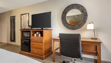 Habitación de hotel con cama, escritorio de madera, silla de oficina negra, televisor, microondas, refrigerador, espejo y lámpara.