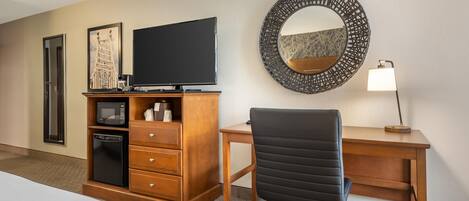 A hotel room with a bed, a wooden desk, a black office chair, a television, a microwave, a refrigerator, a mirror, and a lamp.