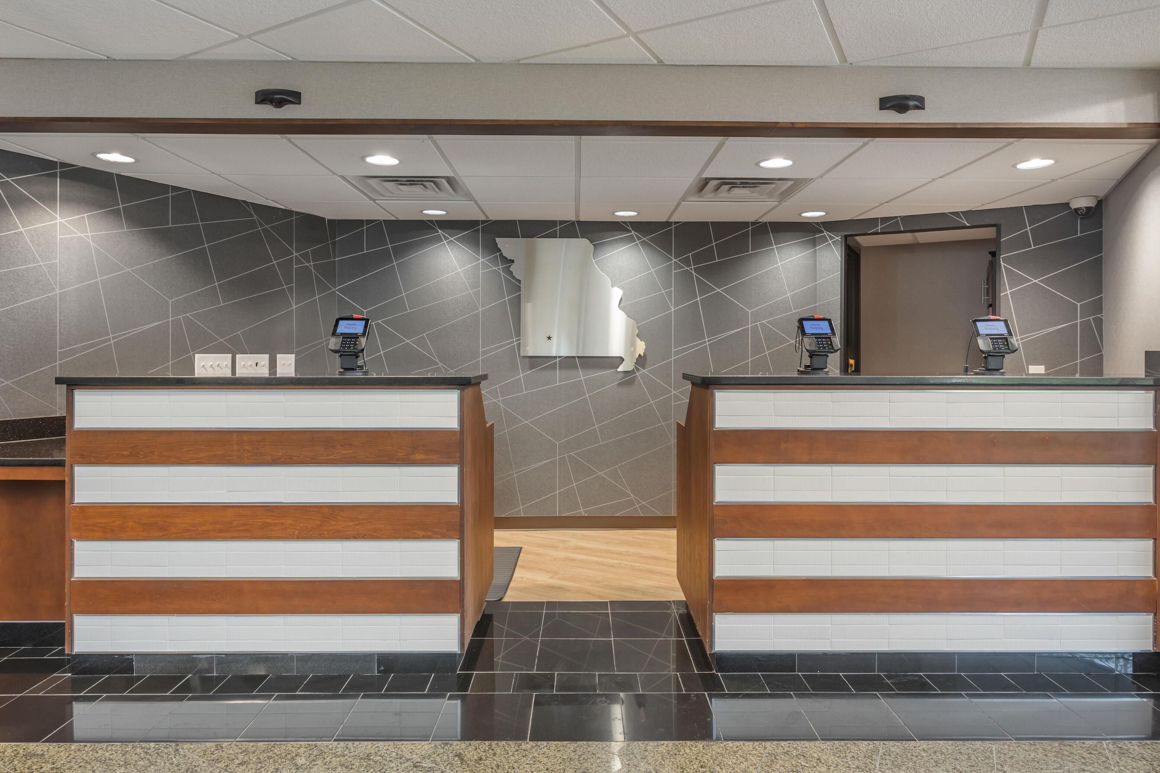A reception area with two counters, each equipped with a digital kiosk. The walls are adorned with a geometric pattern, and the floor is tiled with large, dark tiles.