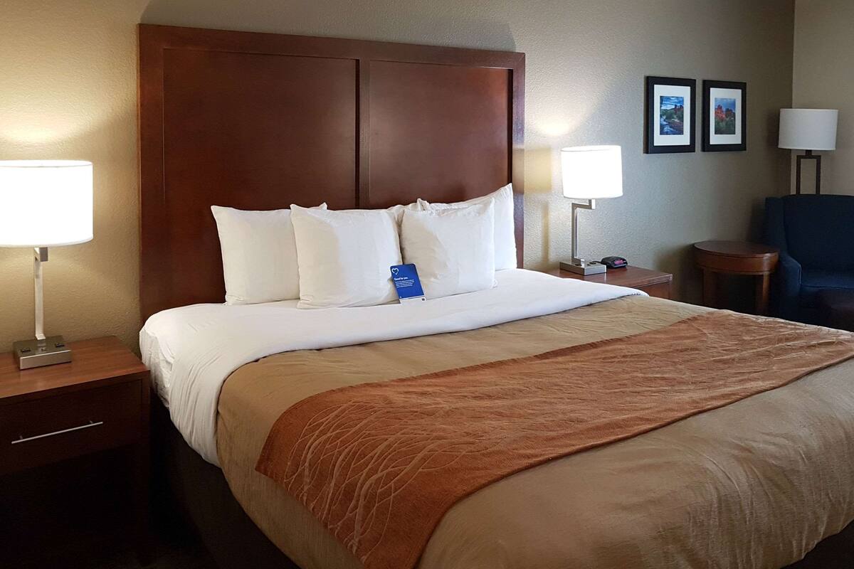Una habitación de hotel con una cama grande, dos lámparas de noche, un cabecero de madera, una manta marrón y dos cuadros en la pared.