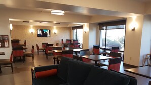 Un espace repas moderne, doté d’un grand canapé, de plusieurs tables et de chaises. Un téléviseur est fixé au mur et de grandes fenêtres laissent entrer la lumière naturelle.
