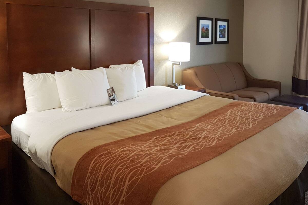 Habitación de hotel con una cama grande, cabecera, mesita de noche con lámpara, una silla y dos cuadros enmarcados en la pared.
