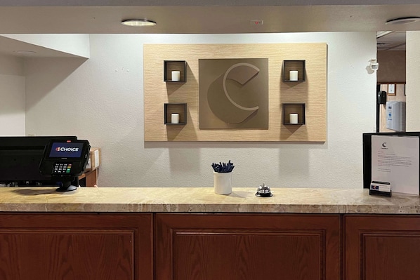A hotel reception area with a wooden wall-mounted sign, a cash register, and a small desk with a cup of pens.