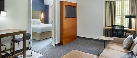 Habitación de hotel con una cama, un escritorio con dos sillas, un sofá, un televisor montado en un panel de madera y una ventana con cortinas.