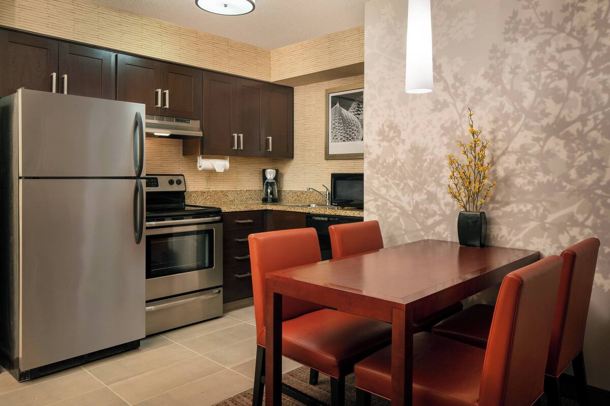 A modern kitchen with dark wooden cabinets, stainless steel appliances, and a dining area with orange chairs.