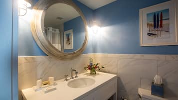 A bathroom with a large round mirror, a sink, and a toilet. There is a framed picture on the wall and a vase with flowers on the sink.