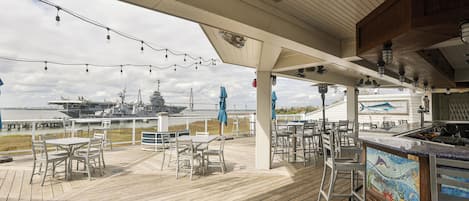 A covered outdoor deck with tables and chairs, overlooking a body of water with a ship in the background.