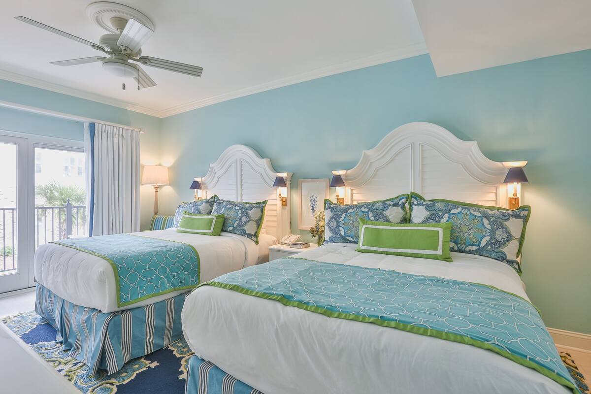 Ein Hotelzimmer mit zwei Betten, jeweils mit weißem Kopfteil und blauer Bettwäsche. Es gibt einen Deckenventilator, ein Fenster mit Vorhängen und einen kleinen Tisch mit einer Lampe.