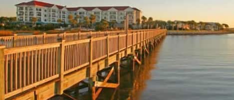 A waterfront hotel with a wooden pier extending into the water, featuring multiple balconies and a red roof.