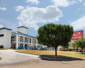 A two-story hotel with a blue roof and white walls, a large tree in the foreground, and a sign for EconoLodge in the background. - Econo Lodge Canton I-55 (Canton)