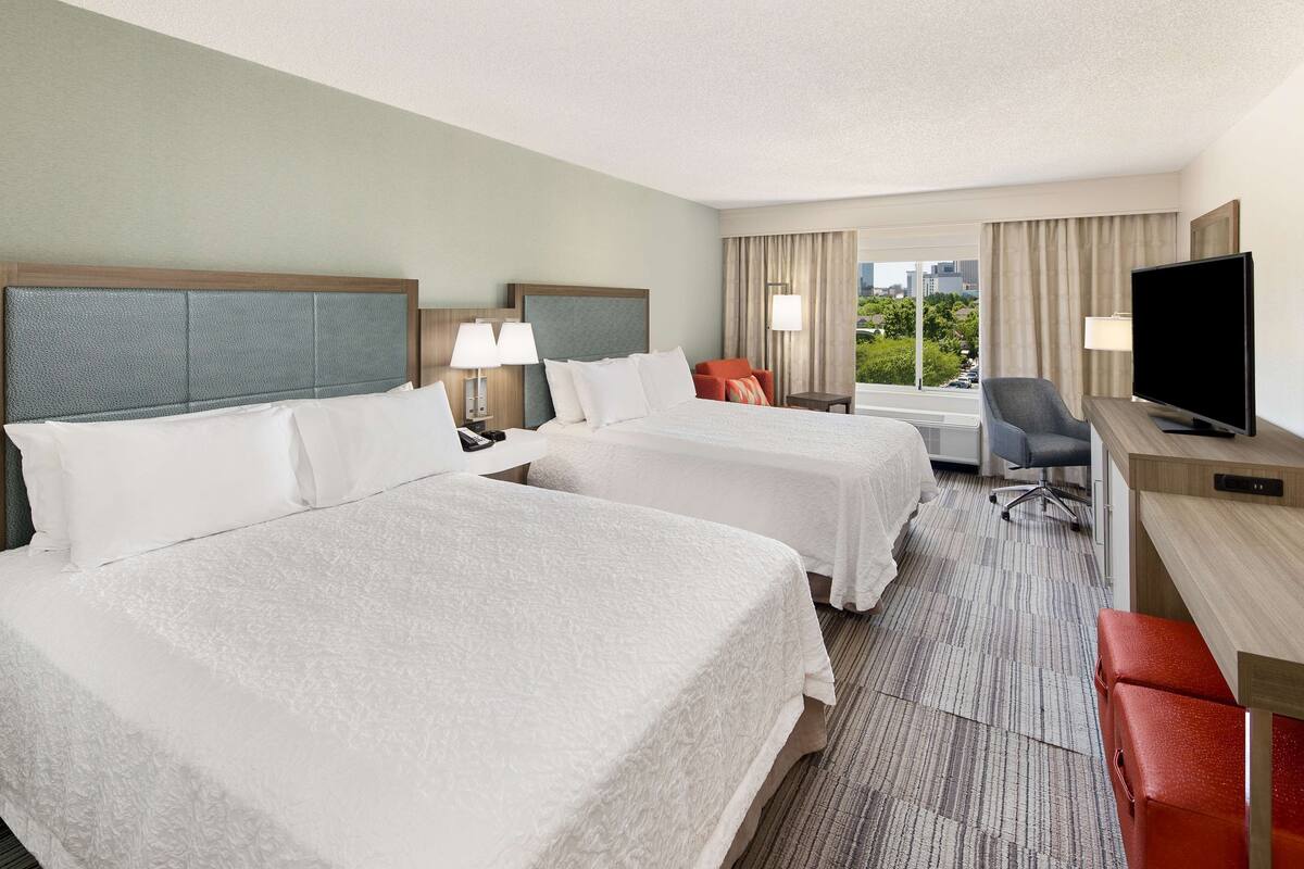 A hotel room with two beds, a desk with a TV, a chair, and a view of the city.