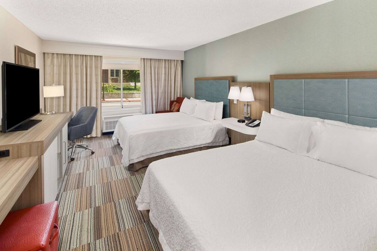 A hotel room with two beds, a desk with a TV, a chair, a desk lamp, a phone, and a window with curtains.