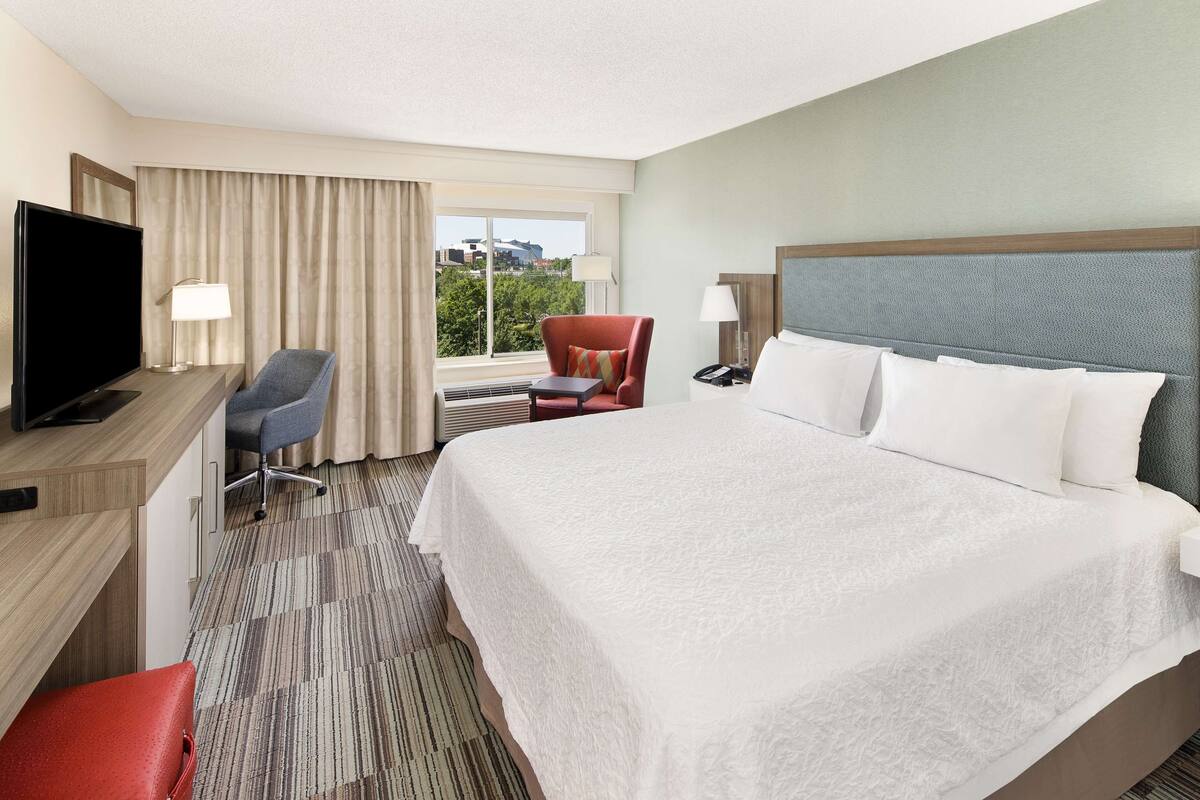 A hotel room with a large bed, a desk with a TV, a chair, a desk lamp, and a view of the outdoors.