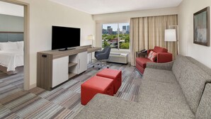 A hotel room with a bed, a TV, a desk, a chair, a sofa, and a window with a city view.
