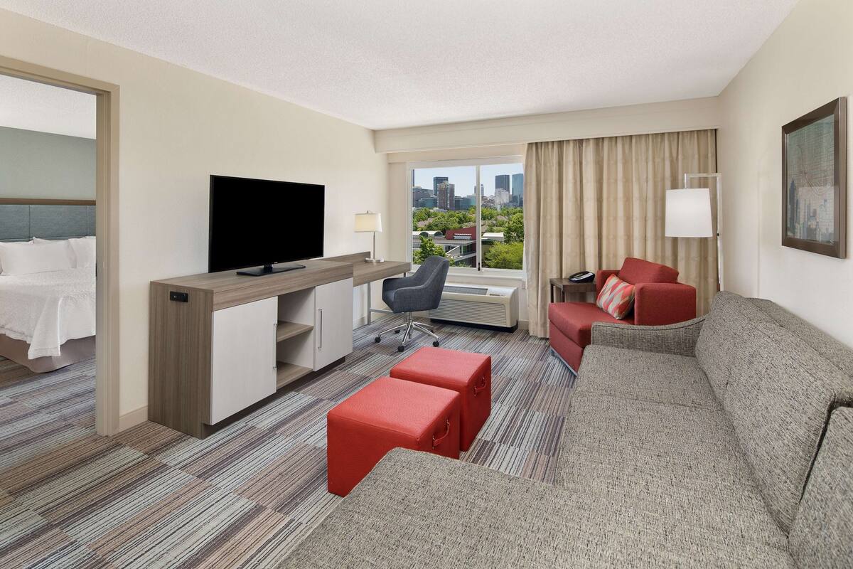 A hotel room with a bed, a TV, a desk, a chair, a sofa, and a window with a city view.
