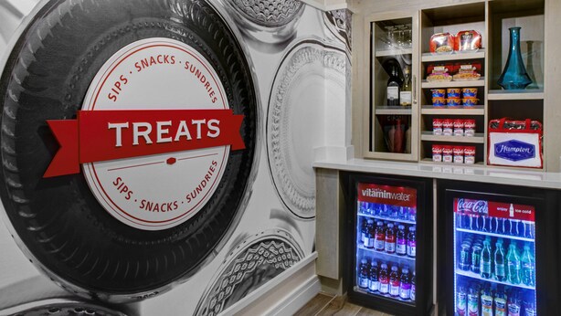 A wall with a large tire design labeled "SIPS, SNACKS, SUNDRIES" and a "TREATS" sign. There are two beverage dispensers below, one with Vitaminwater and the other with Coca-Cola.