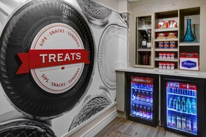 A wall with a large tire design labeled "SIPS, SNACKS, SUNDRIES" and a "TREATS" sign. There are two beverage dispensers below, one with Vitaminwater and the other with Coca-Cola. - Hampton Inn Atlanta-Georgia Tech-Downtown (Atlanta)