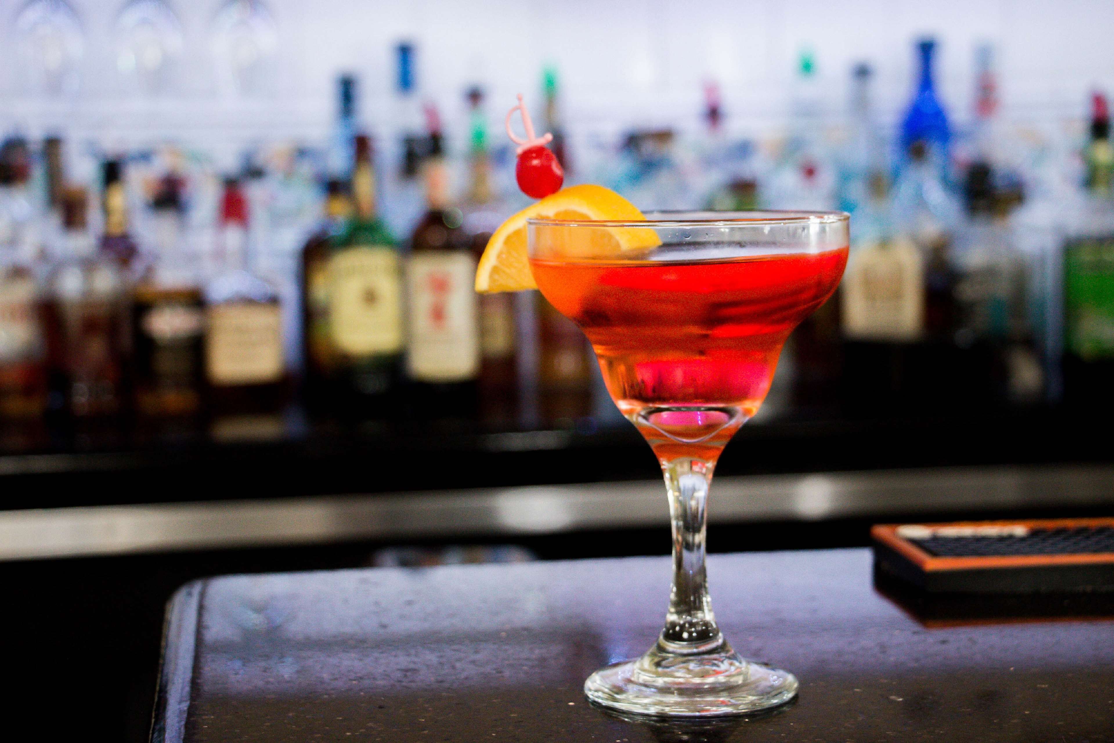 A cocktail in a glass with a cherry and orange slice garnish, set on a bar counter with a blurred background of various bottles.