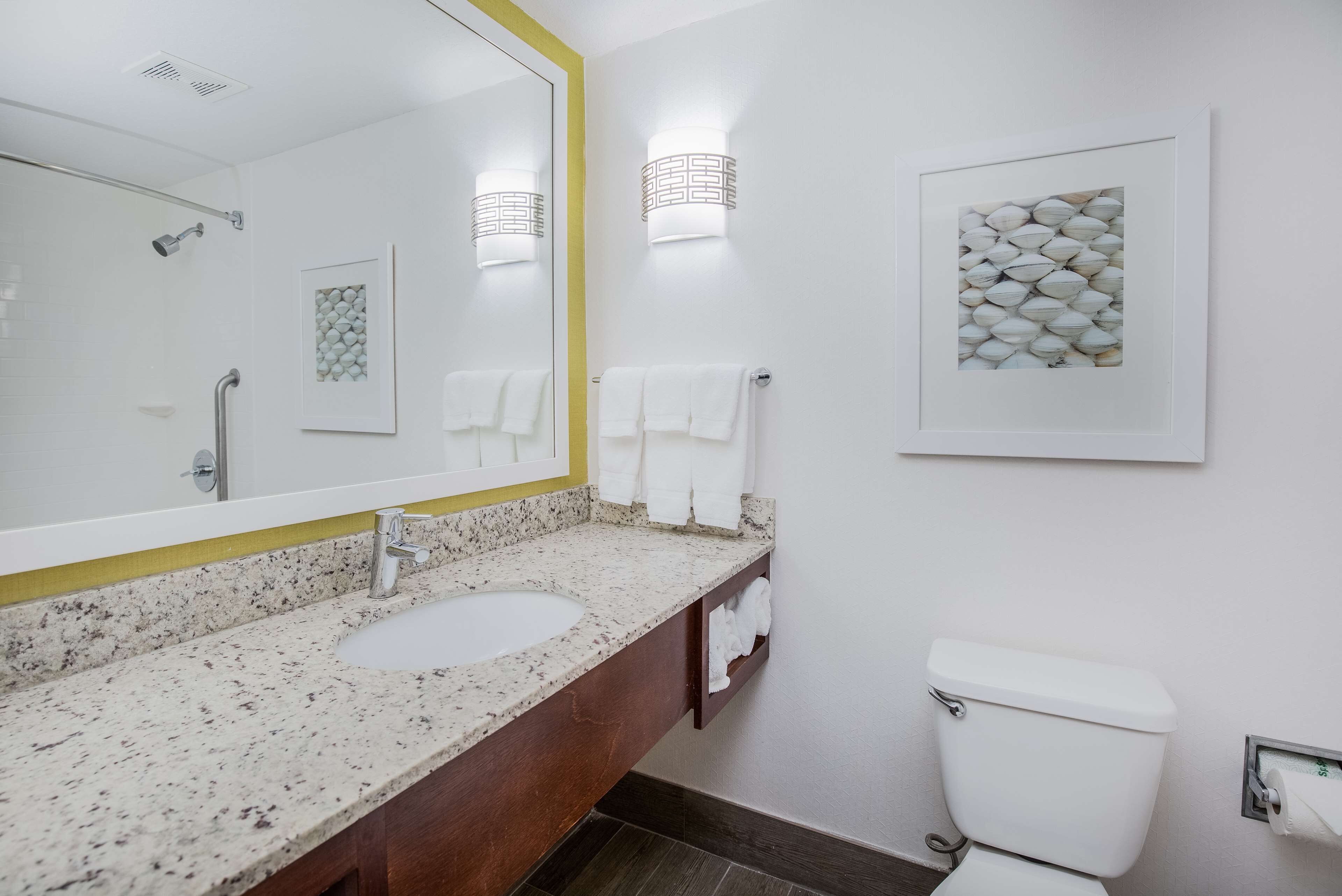 Un baño con lavamanos de encimera de granito, un espejo grande, un inodoro blanco y dos luces empotradas.