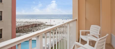 A balcony with a view of the beach, a pool, and white plastic chairs.