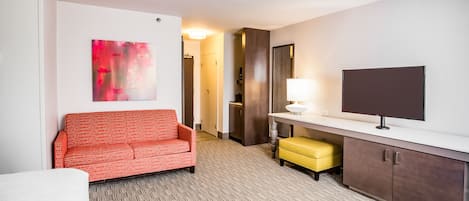 A hotel room with a red patterned sofa, a yellow ottoman, a flat-screen TV, and a painting on the wall.