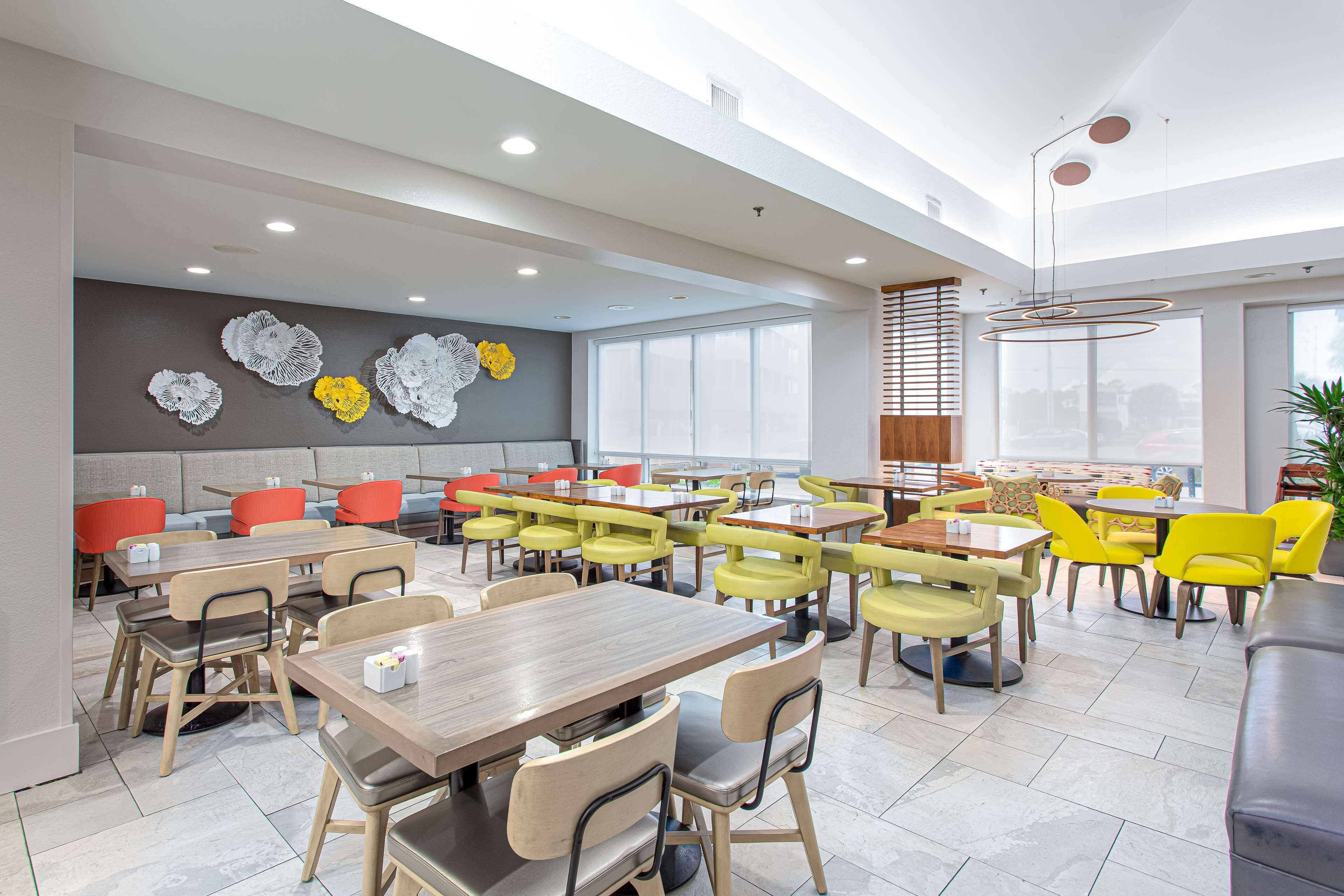 A modern dining area with multiple tables and chairs, a grey wall with decorative elements, and large windows allowing natural light.