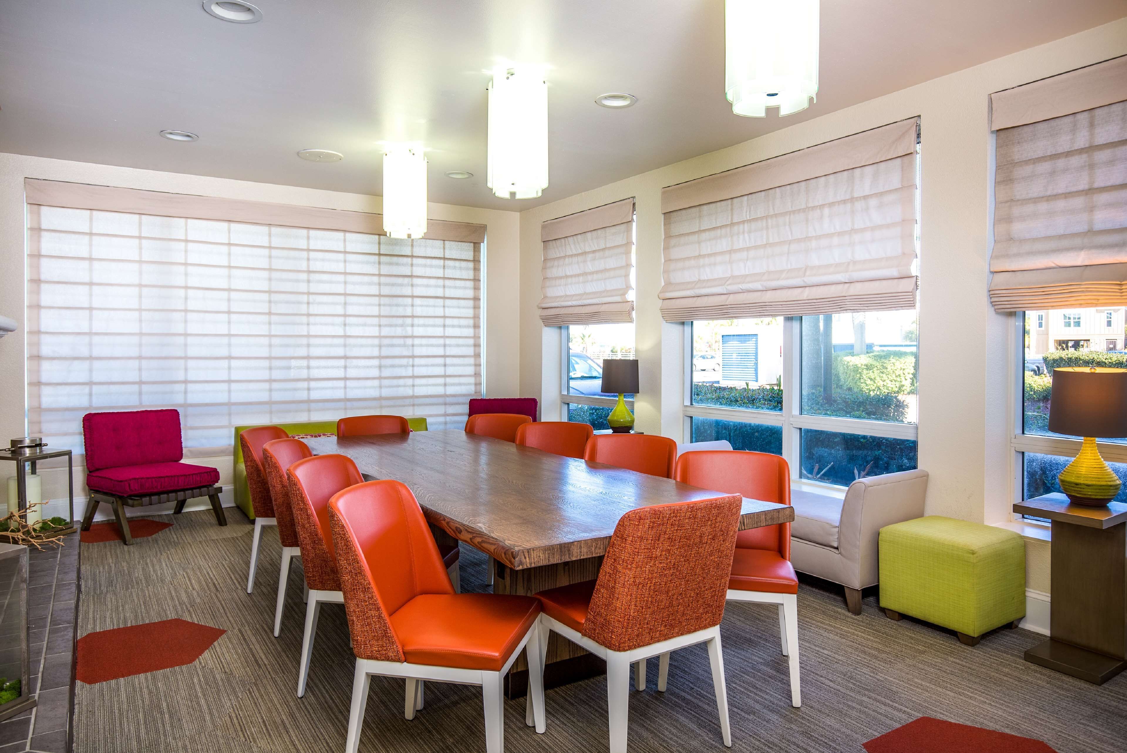 A conference room with a large wooden table surrounded by orange chairs, beige walls, and large windows with blinds.