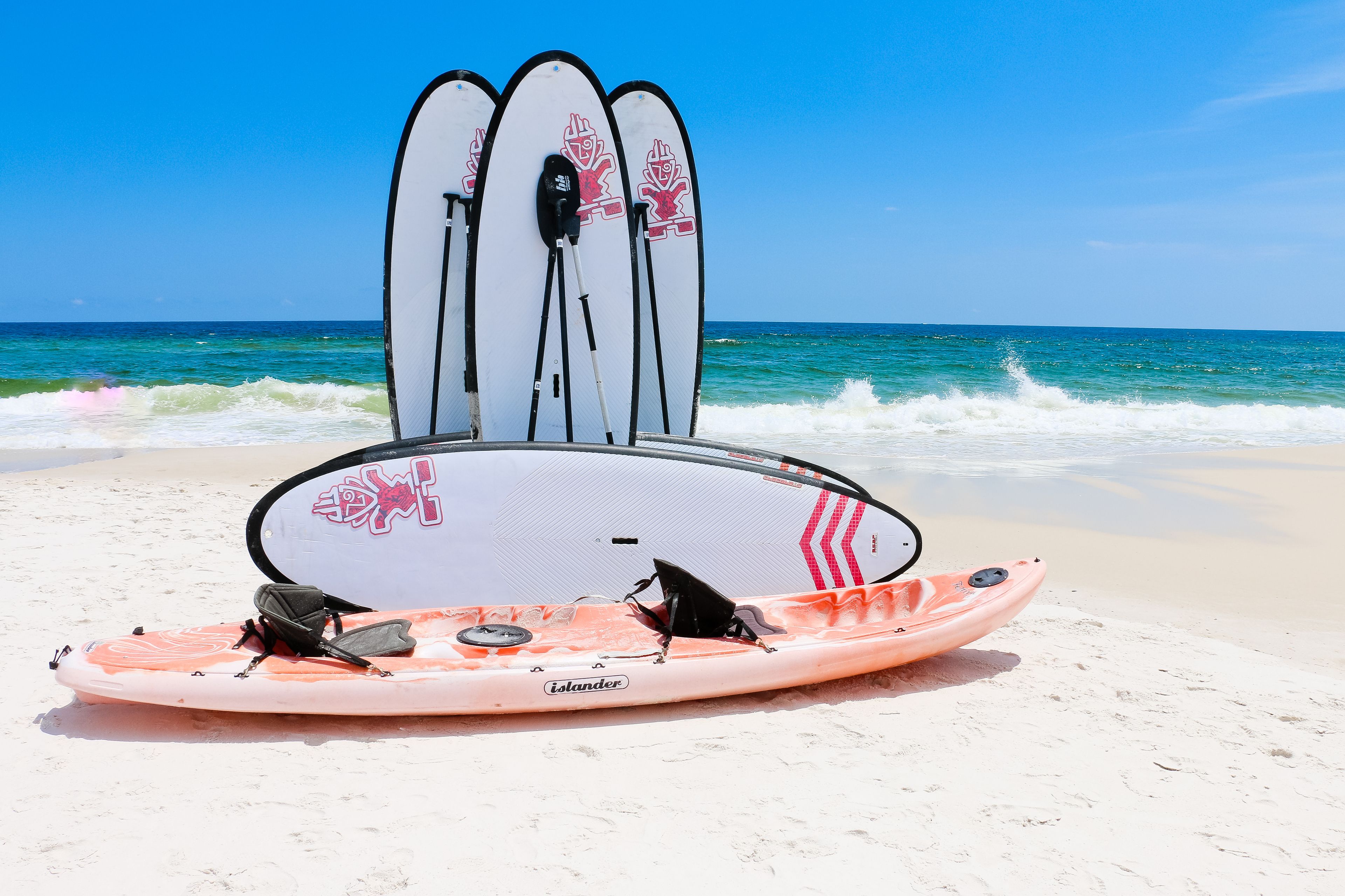 Ein Kajak mit einer schwarzen Tasche an einem sandigen Strand, im Hintergrund Surfbretter.