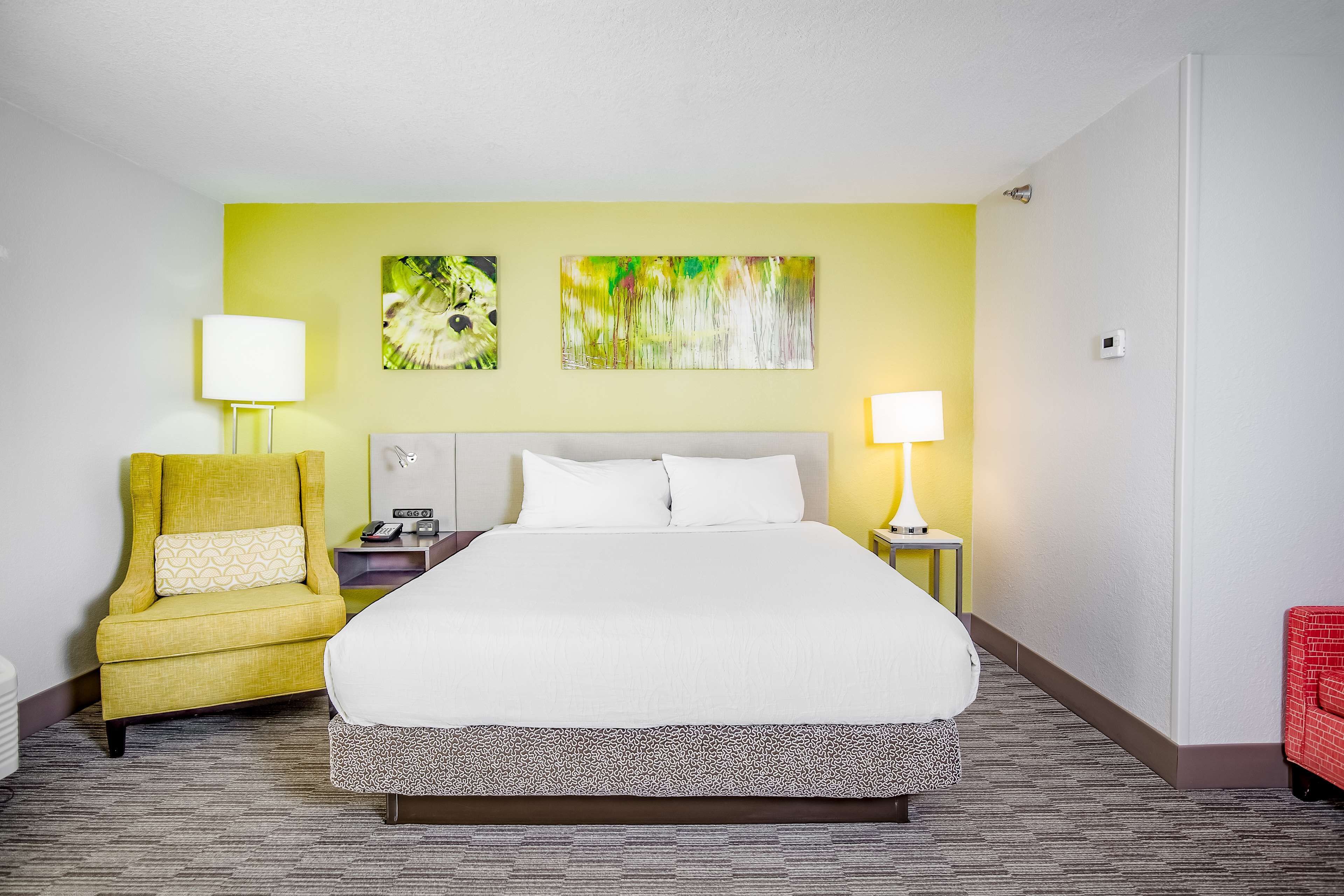 A hotel room with a large bed, a yellow armchair, two wall-mounted paintings, and a red chair.