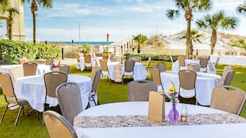 An outdoor dining area with round tables and chairs, set against a backdrop of palm trees and a beach view.