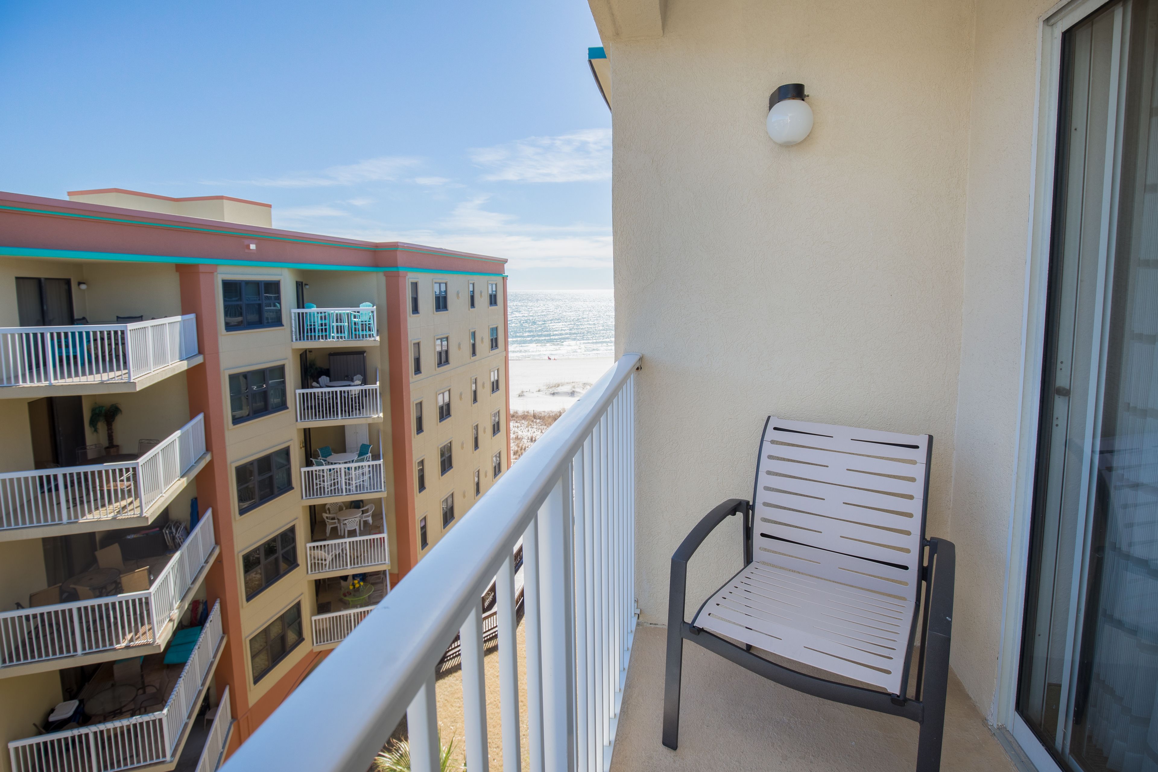 A balcony with a chair, a railing, and a view of the beach and adjacent buildings.