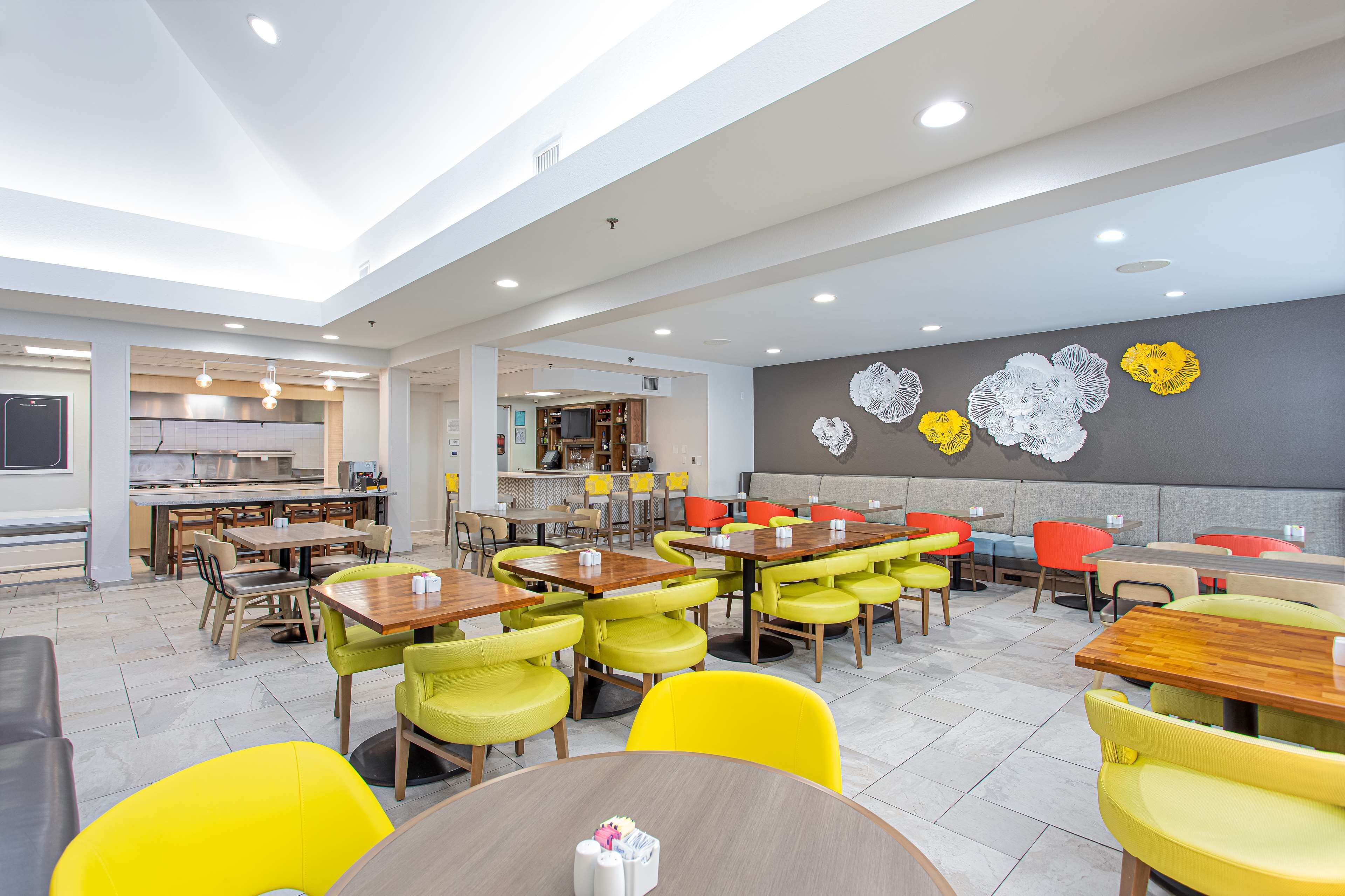 A modern dining area with multiple tables and chairs, a kitchen in the background, and decorative wall art.