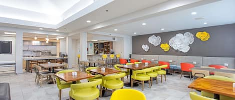 A modern dining area with multiple tables and chairs, a kitchen in the background, and decorative wall art.