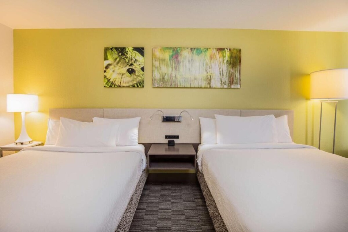 A hotel room with two beds, a headboard, a nightstand, and two wall-mounted lamps.