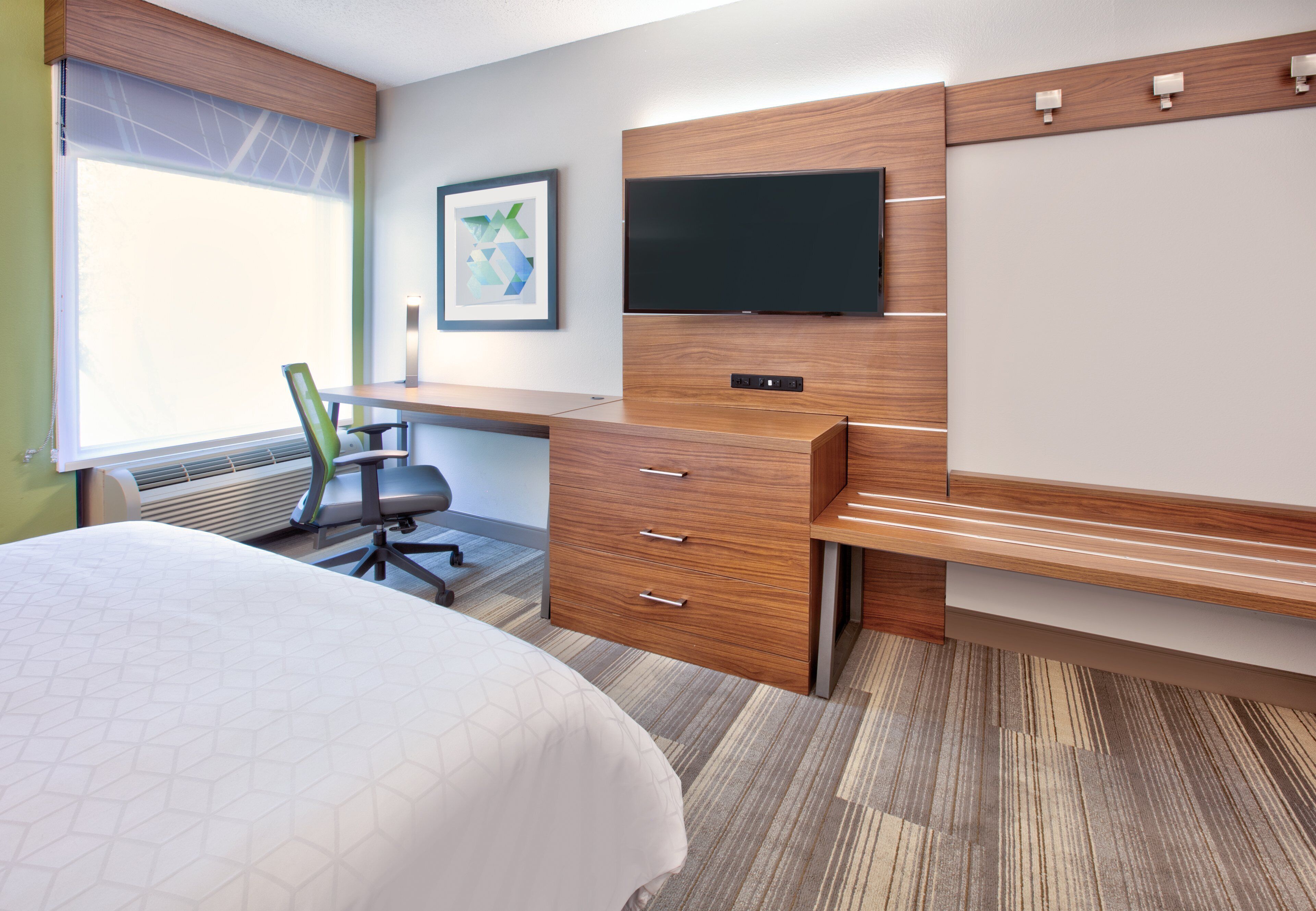 Habitación de hotel con cama, escritorio con silla, televisor de pantalla plana montado en una pared de madera y un ventanal grande.