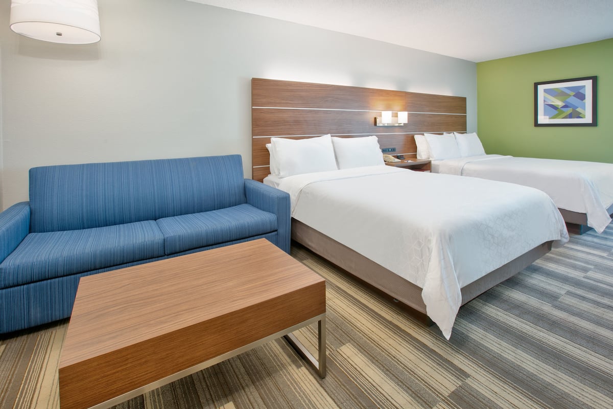 A hotel room with a blue sofa, a wooden coffee table, two beds with white linens, a wooden headboard, a nightstand, and a wall-mounted lamp.