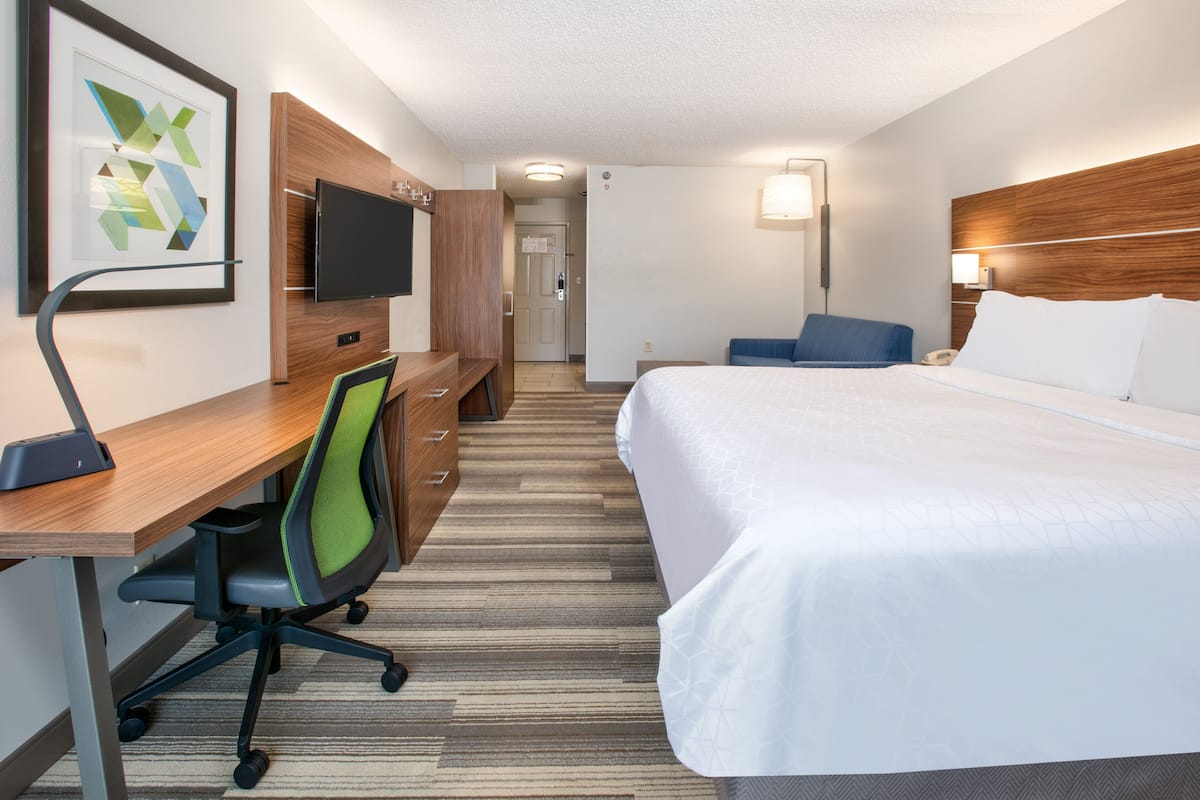 A hotel room with a bed, desk, chair, TV, and a door leading to a hallway.