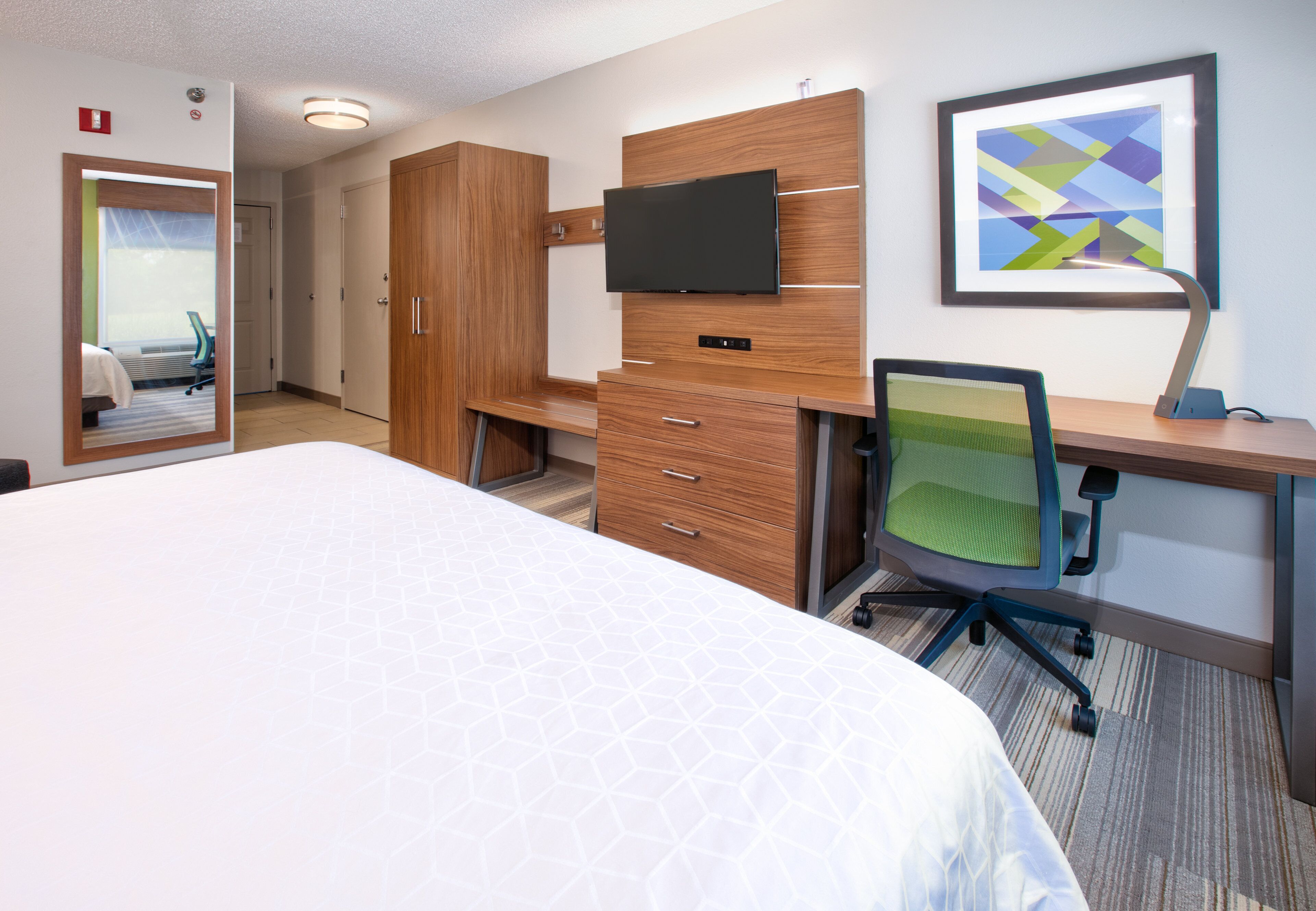Une chambre d’hôtel avec un grand lit, une armoire en bois, un bureau avec un ordinateur, une chaise et un téléviseur à écran plat fixé au mur.