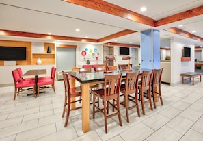 Uma sala de jantar ampla com uma mesa grande e várias cadeiras, área de cozinha com geladeira e pia, e uma televisão fixada na parede.
