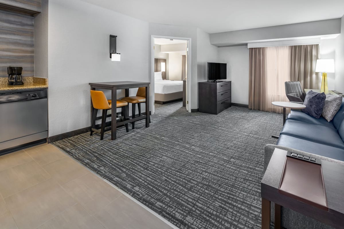 A hotel room with a kitchenette, a dining area with a table and chairs, a bed, a TV, a sofa, and a lamp.
