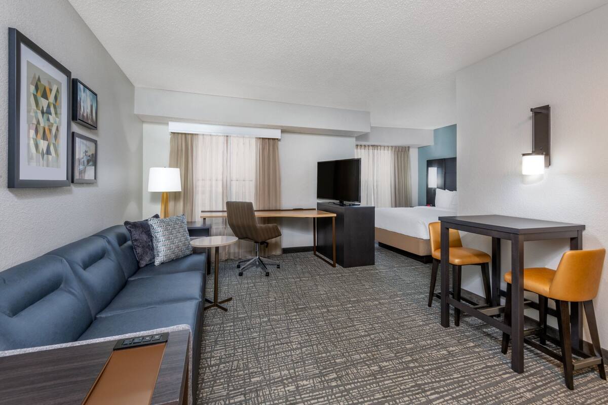 A hotel room with a blue sofa, a desk with a chair, a bed, a TV, and a small table with two chairs.