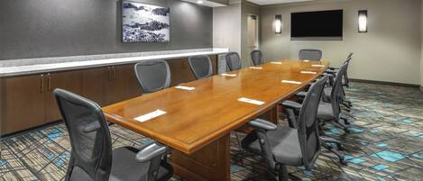 A conference room with a long wooden table, black office chairs, a large flat-screen TV, and a patterned carpet.