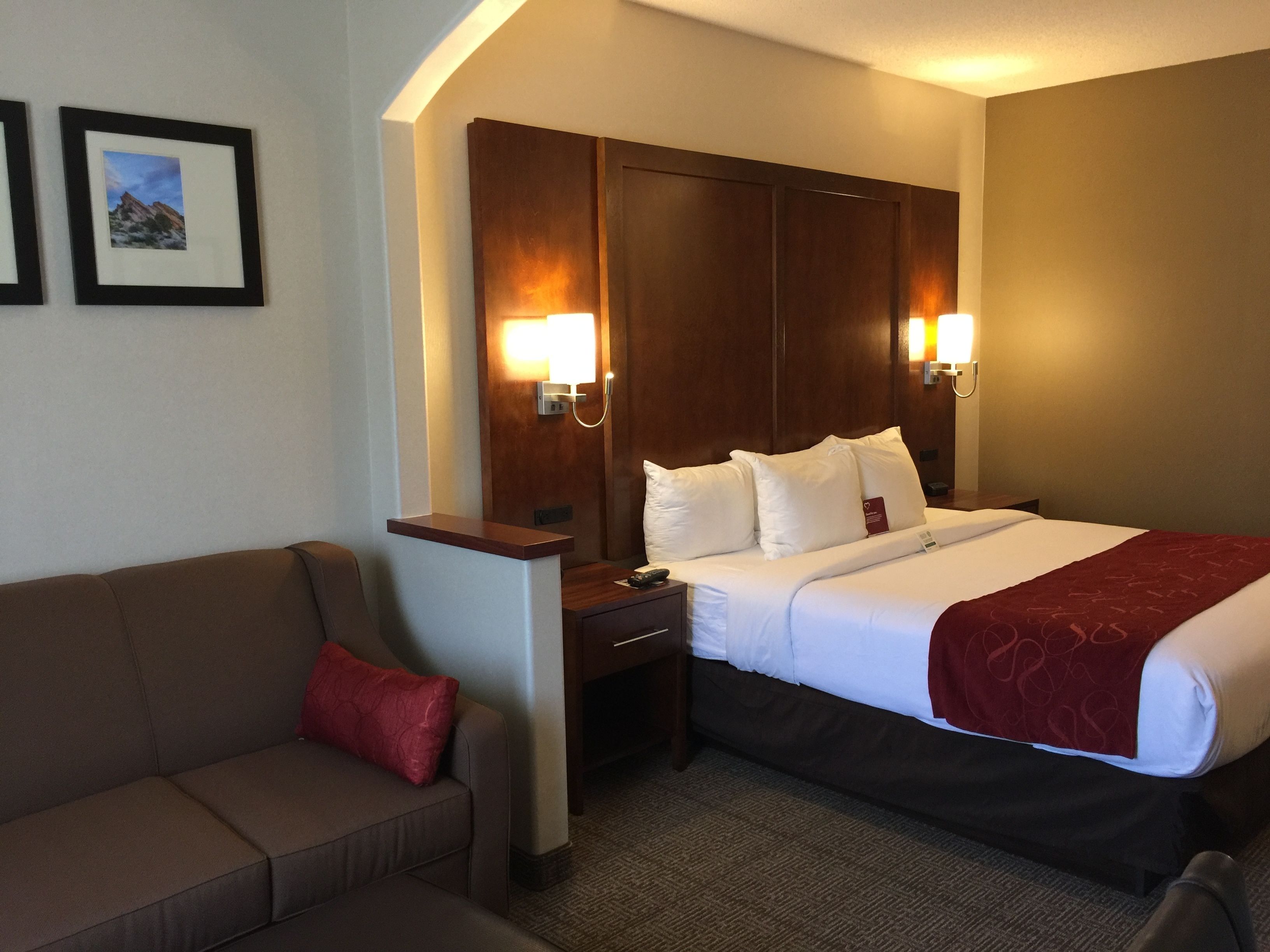 A hotel room with a bed, a wooden headboard, a nightstand, a red throw blanket, a brown sofa, and two wall-mounted lamps.