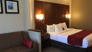 A hotel room with a bed, a wooden headboard, a nightstand, a red throw blanket, a brown sofa, and two wall-mounted lamps.