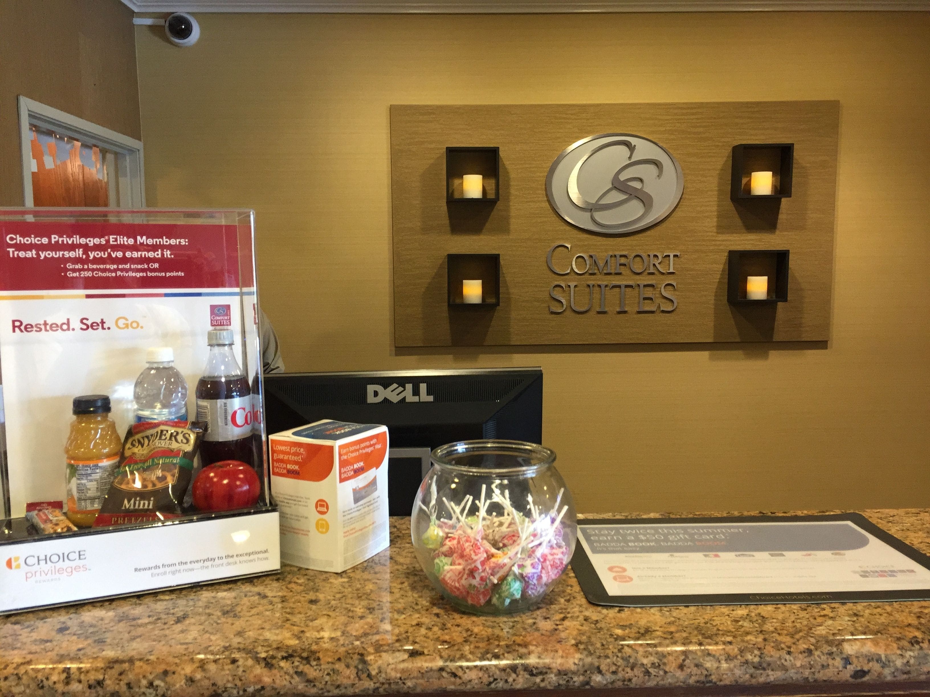 A hotel reception area with a sign for 'Comfort Suites', a Dell computer, and a display of snacks and beverages.