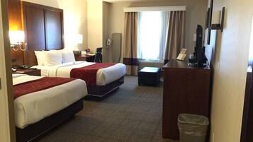 A hotel room with two beds, a desk, a chair, a trash bin, and a large window with curtains.