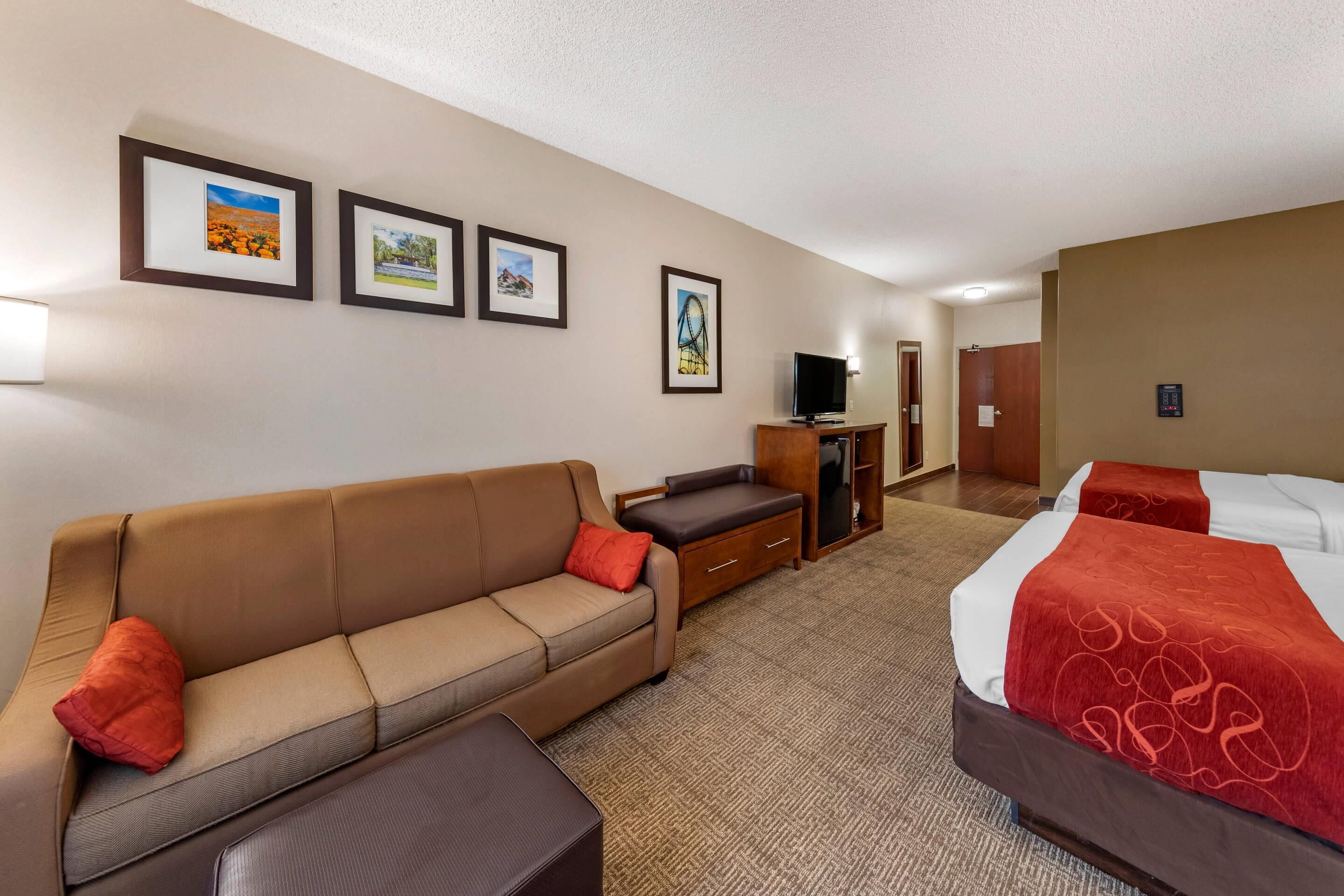 A hotel room with a brown sofa, a small desk, a TV, and two beds with red blankets. There are framed pictures on the wall.